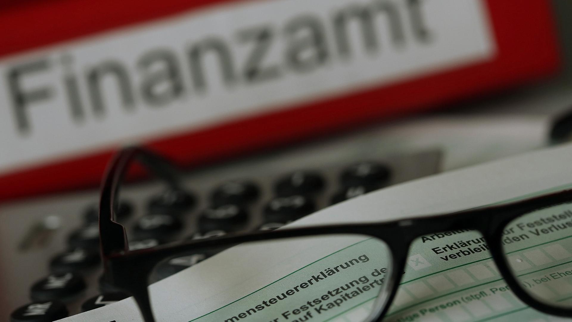 Auf einem Vordruck für die Einkommensteuererklärung liegen vor einem Aktenordner mit der Aufschrift "Finanzamt" eine Brille und ein Kugelschreiber