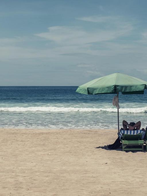 Bild eines fast komplett leeren Strands mit Blick aufs Meer: Nur zwei Menschen liegen auf ihren Strandliegen unter einem Sonnenschirm.