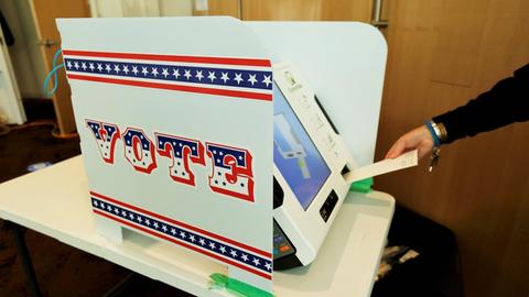 Auf einem Tisch steht eine Wahlmaschine mit einem Sichtschutz, auf dem die Aufschrift "Vote" zu lesen ist.