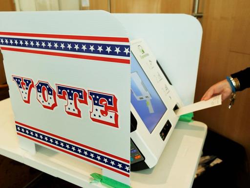 Auf einem Tisch steht eine Wahlmaschine mit einem Sichtschutz, auf dem die Aufschrift "Vote" zu lesen ist.