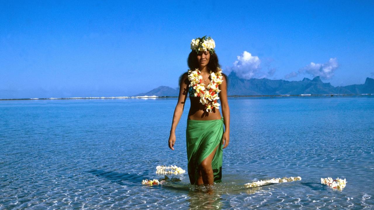 Eine junge Frau aus Tahiti mit Blütenketten geschmückt und einem grünen Pareo um die Hüften geht durch seichtes Wasser.