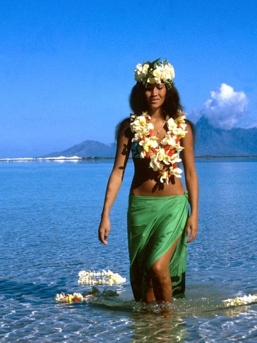 Eine junge Frau aus Tahiti mit Blütenketten geschmückt und einem grünen Pareo um die Hüften geht durch seichtes Wasser.