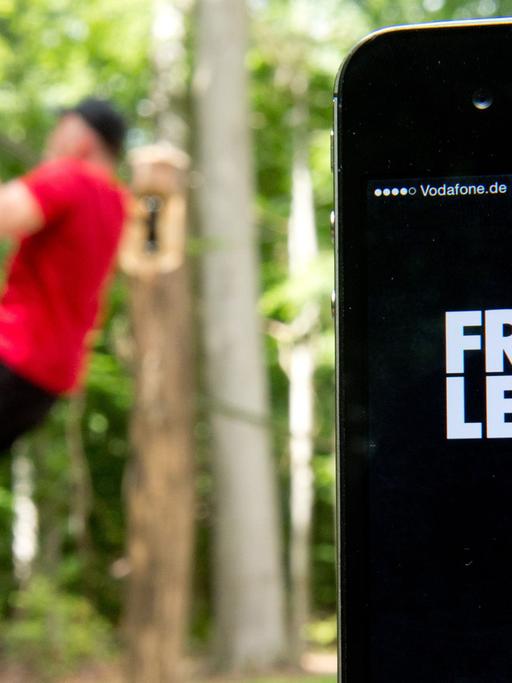 Die Fitness-App "Freeletics" wird auf einem iPhone angezeigt, während im Hintergrund ein Mann auf einem Trimm-Dich-Pfad einen Klimmzug macht.
