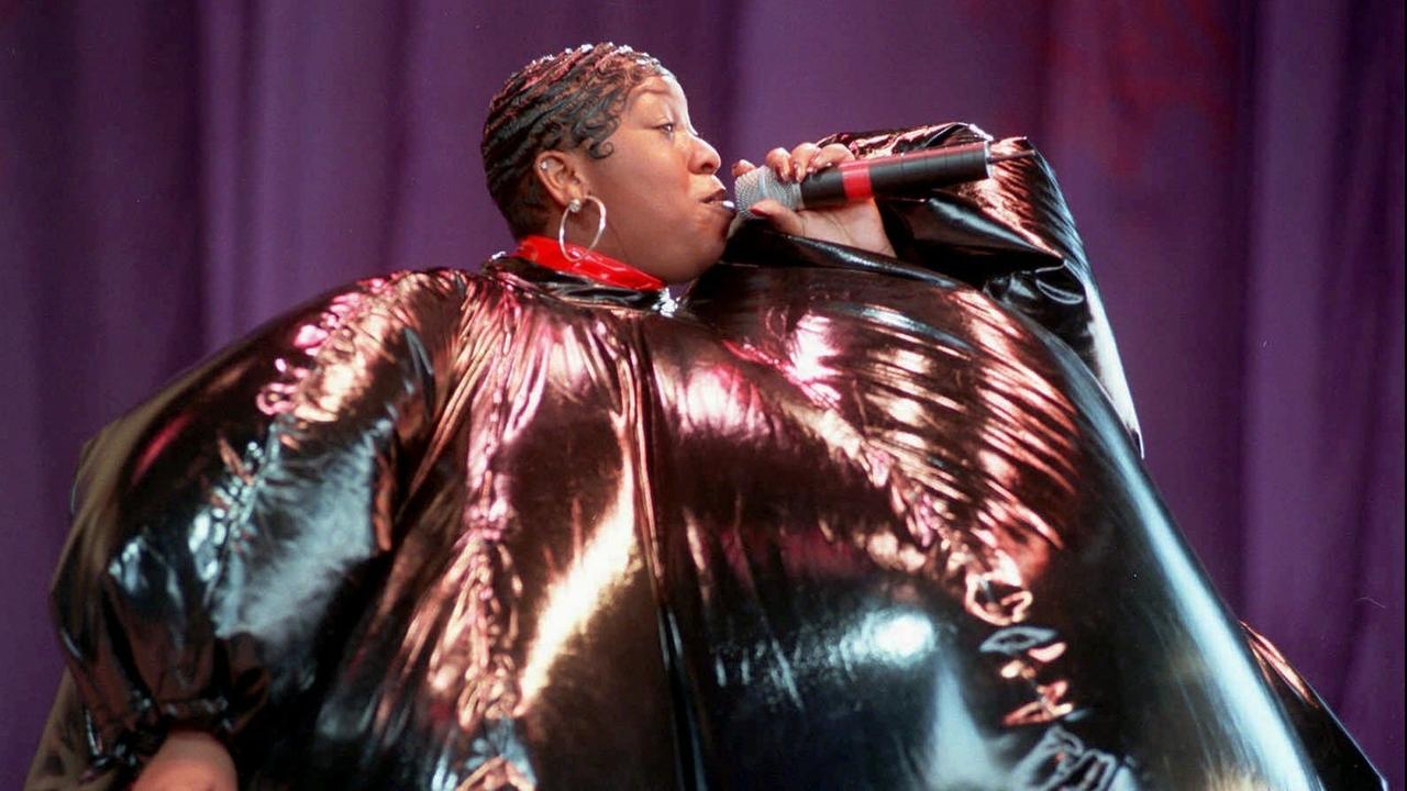 Eine gestylte schwarze Frau mit einem Mikrofon in der Hand. SIe trägt ein überdimensionales schwarz-pinkes Kostüm, das aussieht wie ein aufgeblasener Ballon.