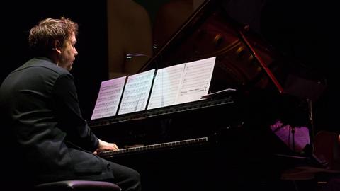 Ein Mann sitzt mit dem Rücken zur Kamera an einem aufgeklappten Flügel. Er blickt beim Spielen auf die Notenblätter vor ihm.