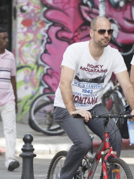 Eine alltägliche Szene aus Tel Aviv: Ein Mann in weißem Hemd und mit Sonnenbrille fährt auf einem Fahrrad durch die Straßen von Tel Aviv. Auf dem Gehweg laufen junge Frauen und ein dunkelhäutiger Mann. Die Häuser sind bunt gestaltet und betonen den alltäglichen Charakter der Szenerie.