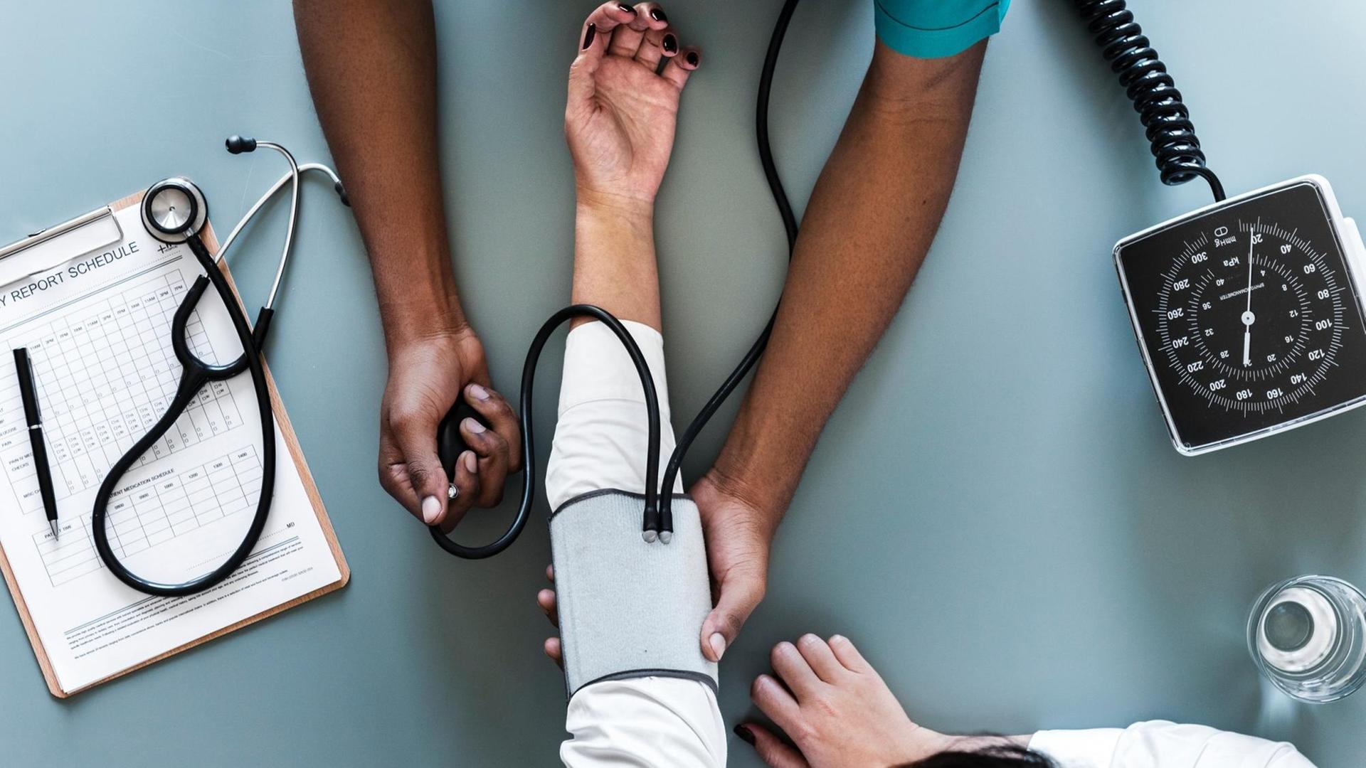 Eine Pflegekraft misst den Blutdruck eines Patienten. Von beiden sind nur die Hände und Arme zu sehen.