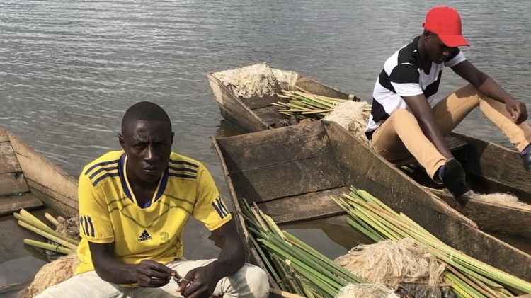 Zwei junge afrikanische Männer sitzen jeweils in einem Holzboot. Einer trägt ein gelbes Fußballtrikot als T-Shirt, der andere eine rote Baseballkappe.