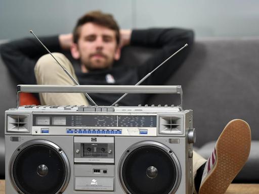 Ein dunkelhaariger Mann sitzt auf einem grauen Sofa und hat einen Fuß auf dem Couchtisch abgelegt. Vor ihm ein altmodischer Kassettenrekorder.