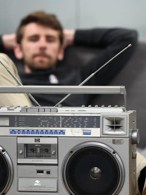 Ein dunkelhaariger Mann sitzt auf einem grauen Sofa und hat einen Fuß auf dem Couchtisch abgelegt. Vor ihm ein altmodischer Kassettenrekorder. Ein dunkelhaariger Mann sitzt auf einem grauen Sofa und hat einen Fuß auf dem Couchtisch abgelegt. Vor ihm ein altmodischer Kassettenrekorder.
