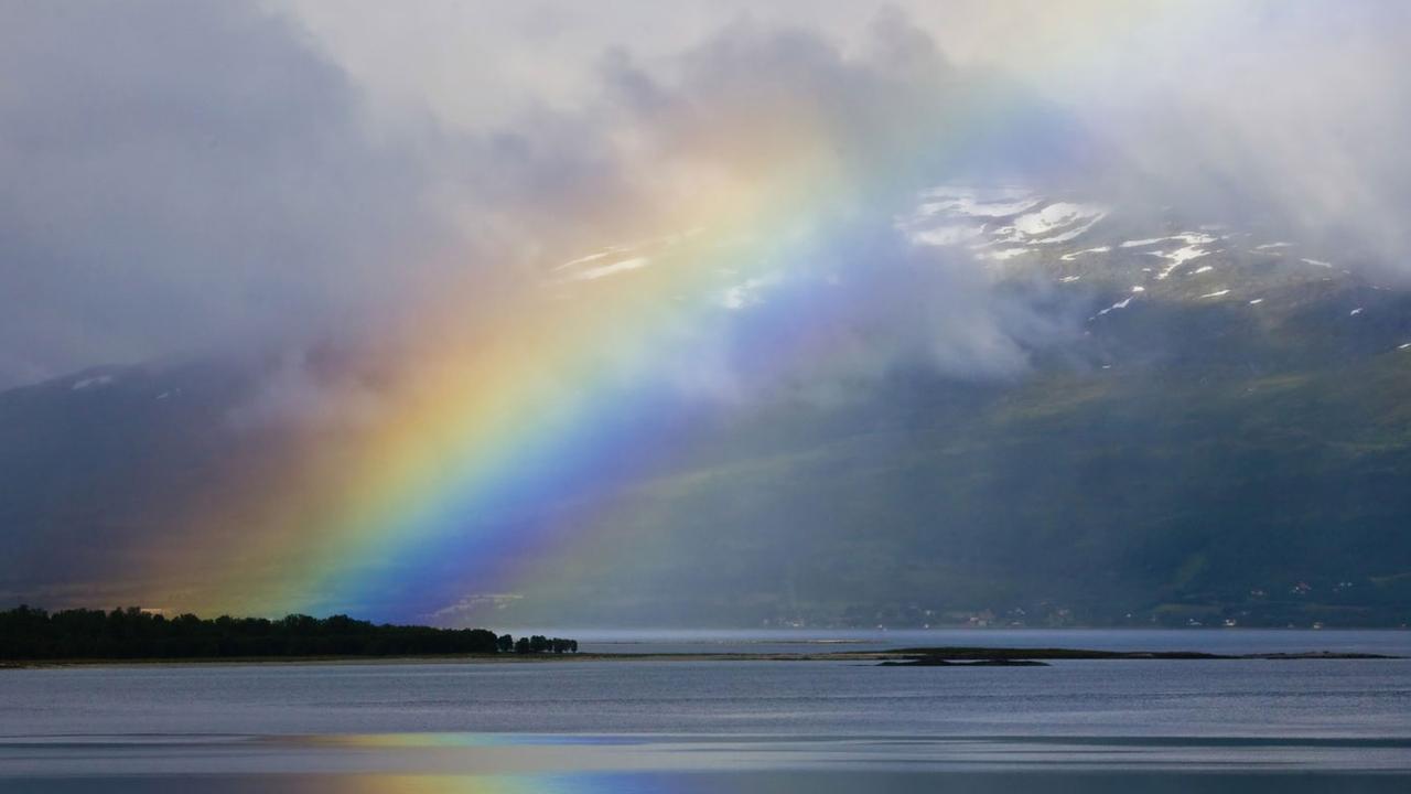 Ein farbenprächtiger Regenbogen, teilweise von niedrig hängenden Wolken verdeckt, spiegelt sich im Wasser vor der Küste der Insel Kvalöya.