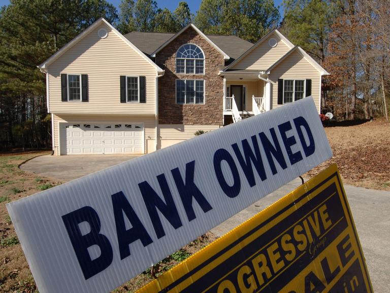 Ein mit Holz verkleidetes Haus in den USA, im Vordergrund ein Schild, auf dem "Bank owned" steht.