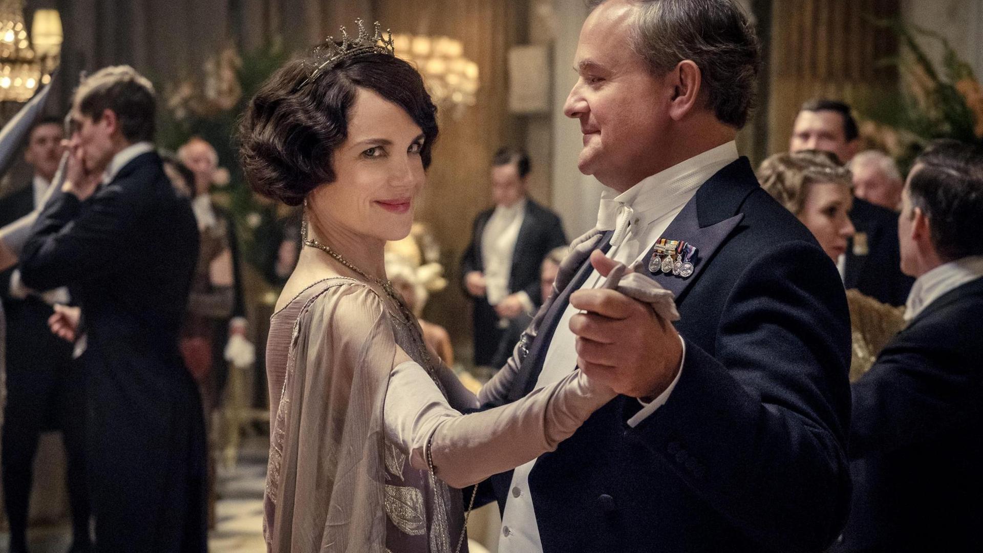 Szenenausschnitt aus dem Film "Downton Abbey" nach der gleichnamigen Fernsehserie, in dem Lord und Lady Grantham, dargestellt durch Hugh Bonneville und Elizabeth McGovern, elegant gekleidet beim Tanzen zu sehen sind.