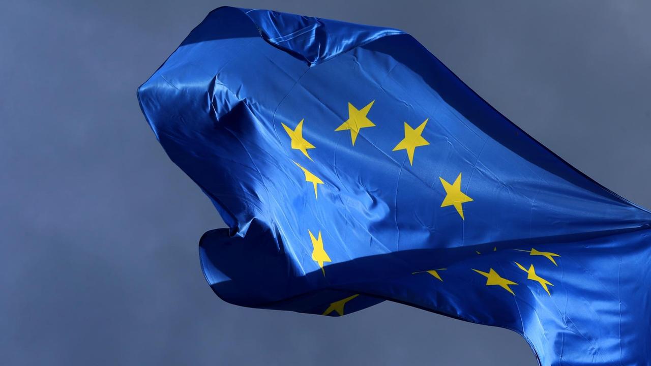 Die sonenbeschienene blaue Flagge mit dem gelben Sternenkreis flattert vor einem dunklen Himmel. 