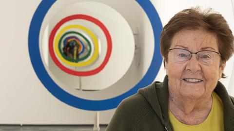Eine ältere Frau mit kurzen Haaren steht in einem Museum vor einem Kunstwerk und lächelt in die Kamera.
