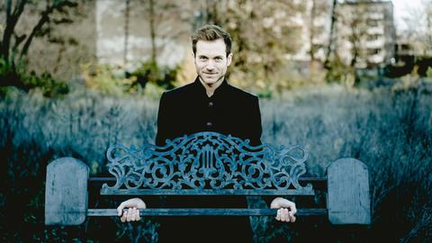 Ein Mann hält das Notenpult von einem alten Flügel vor sich. Er steht dabei im Grünen, weit im Hintergrund sind Häuser unscharf zu erkennen.