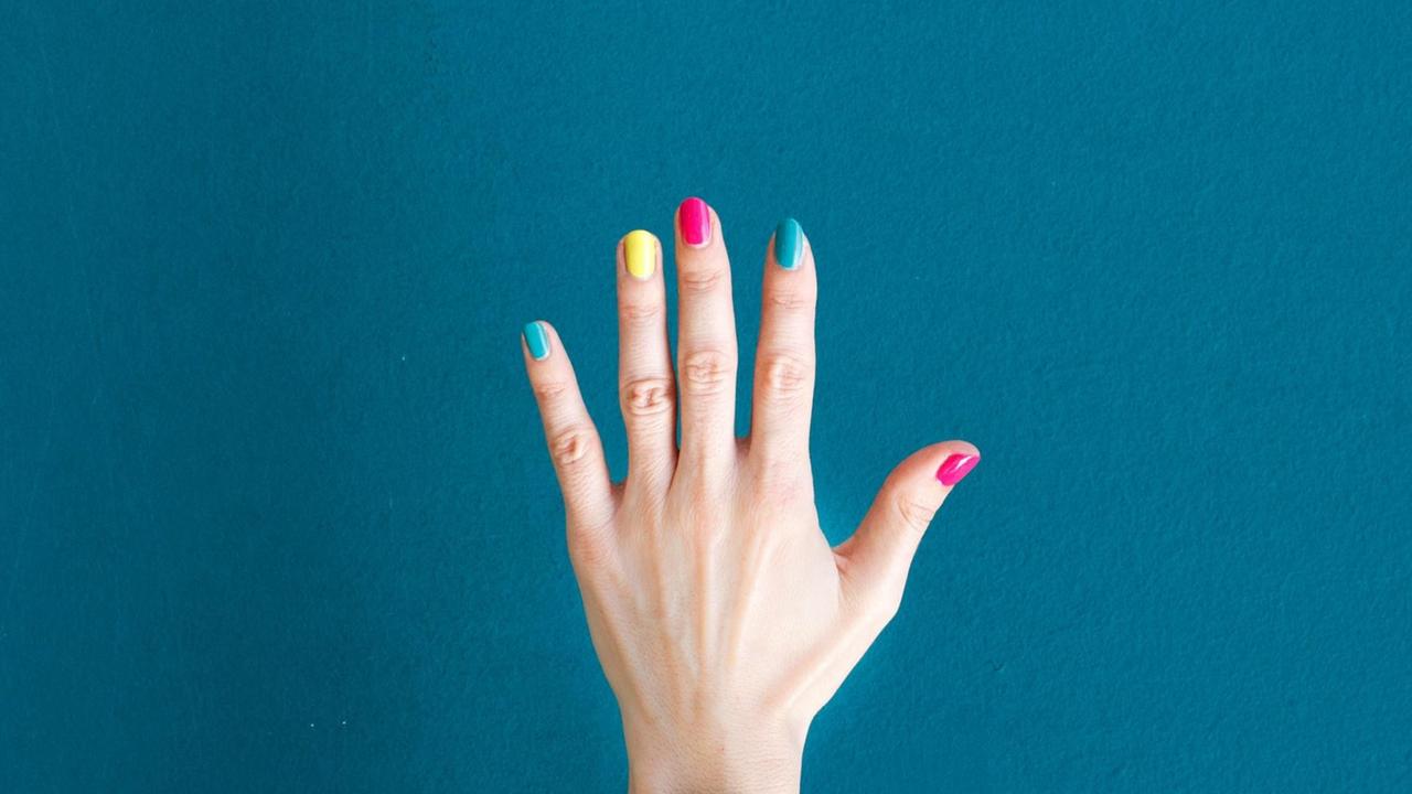 Eine Hand mit buntlackierten Fingernägeln vor blauem Hintergrund.