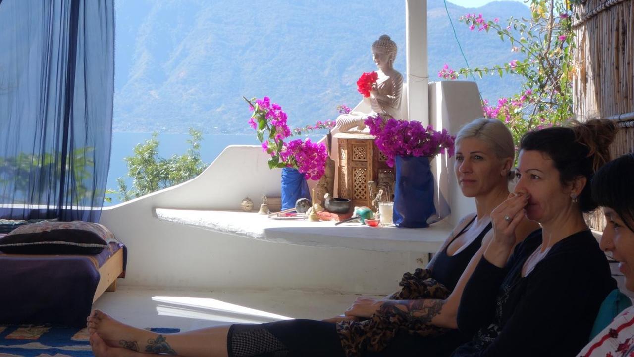 Yogalehrer und Reikimeister sitzen auf einer Terrasse am und nehmen am "Mayan Abdominal Massage"-Kurs teil. Im Hinterggrund ist der Vulkan Atitlan in Guatemala zu sehen.