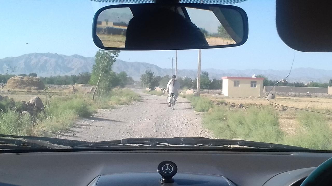 Auf dem Weg zum Safran-Garten: Blick aus dem fahrenden Auto auf eine staubige Schoterpiste, vor dem Wagen ein Fahrradfahrer im langen weißen Gewand.