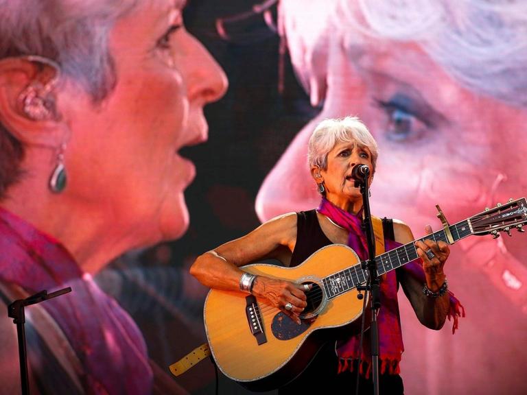 Eine Sängerin mit kurzem, grauen Haar steht mit ihrer Gitarre auf der Bühne, während ihr Porträt zwei Mal übergroß auf Leinwänden hinter ihr erscheint.