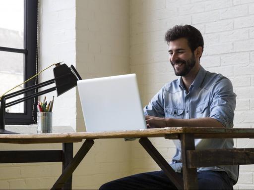Ein lächelnder Mann sitzt an einem Schreibtisch, vor ihm steht ein Laptop
