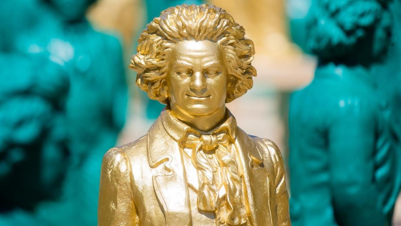 Lächelnde Beethoven-Statuen, ein Kunstwerk des Konzeptkünstlers und Bildhauers Ottmar Hörl, stehen auf dem Münsterplatzin Bonn. Zentral im Bild eine goldene Statue, im Hintergund mehrere türkise.