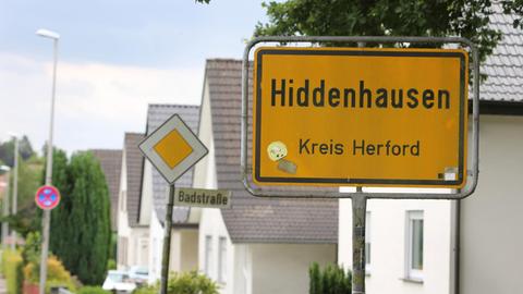 Das Bild vom 08.07.2015 zeigt das Ortseingangsschild von Hiddenhausen (Kreis Herford) an einer Strasse mit Wohnhäusern, die älter als zwanzig Jahre sind und zum Teil renoviert wurden.