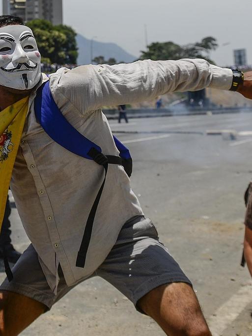 Ein Demonstrant, der eine Guy-Fawkes-Maske trägt und eine venezolanische Flagge um den Hals hat, wirft während einer Auseinandersetzung mit Sicherheitskräften Steine.
