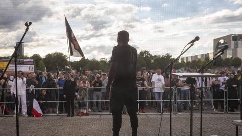 Rückansicht eines Mannes, er steht bei einer Kundgebung auf der Bühne und spricht in ein Mikrofon.
