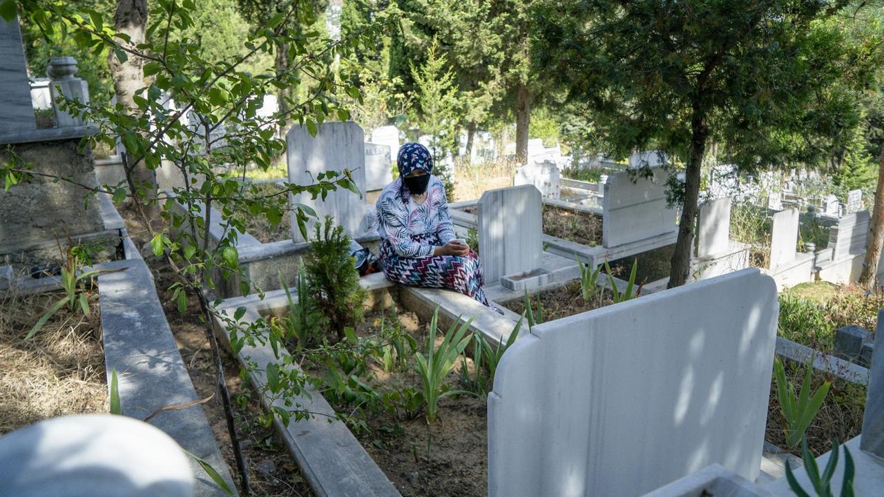 Auf einem grünen Friedhof, sitzt eine ältere Frau am Rand eines Grabs.