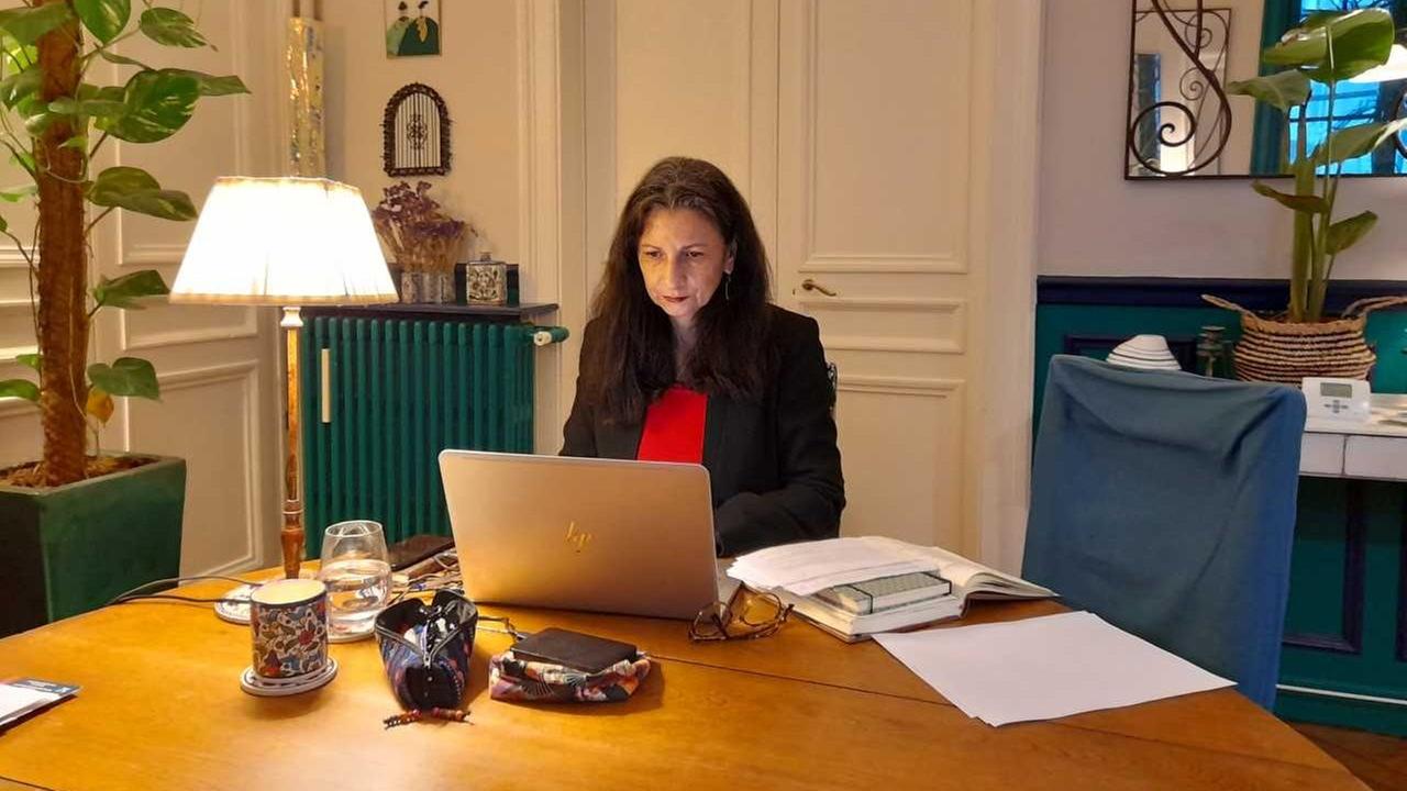 Eine Frau mit langen, dunklen Haaren sitzt über ein Laptop gebeugt an einem großen Tisch aus hellem Holz.