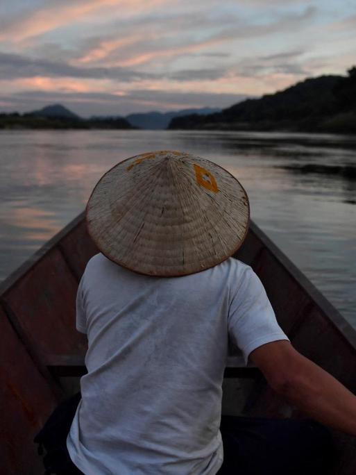 Ein Mann sitzt in einem Fischerboot und fährt in der Dämmerung den Mekong Strom entlang.