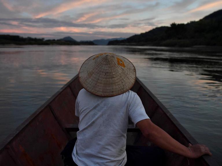Ein Mann sitzt in einem Fischerboot und fährt in der Dämmerung den Mekong Strom entlang.