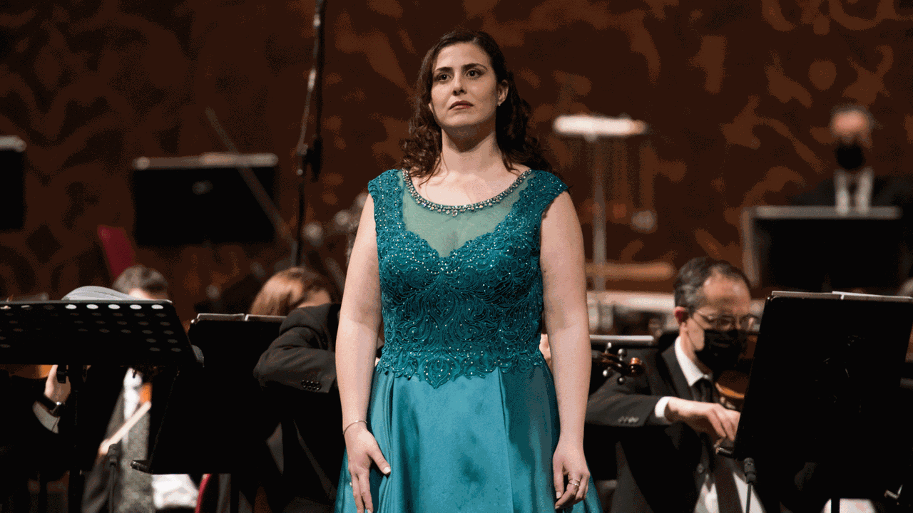 Eine Frau in petrolfarbenem Abendkleid steht vor einem Orchester und schaut ernst in das Publikum vor ihr.
