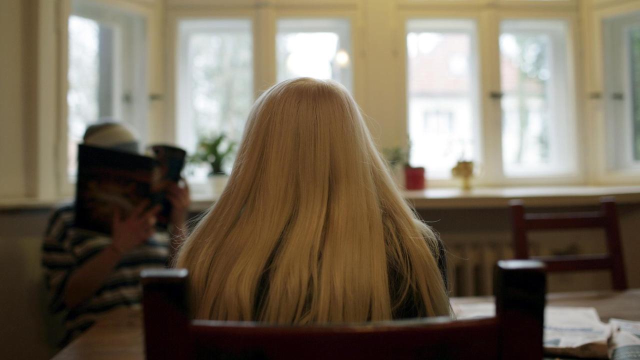 Eine Frau mit langen blonden Haaren sitzt in einem großen Raum mit hohen Fenstern.