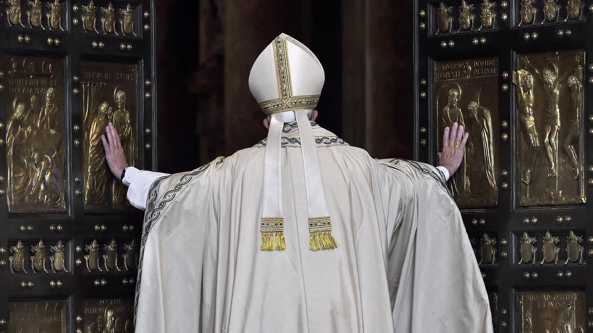Papst Franziskus von Hinten, wie er die großen Flügel der "Heiligen Pforte" öffnet. Das Portal besteht aus zwei großen Kassettentüren mit schwarzen Rahmen und goldenen Kassetten mit Motiven der Heilsgeschichte.