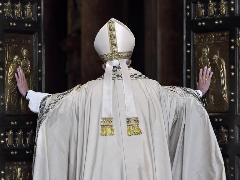 Papst Franziskus von Hinten, wie er die großen Flügel der "Heiligen Pforte" öffnet. Das Portal besteht aus zwei großen Kassettentüren mit schwarzen Rahmen und goldenen Kassetten mit christlichen Motiven.