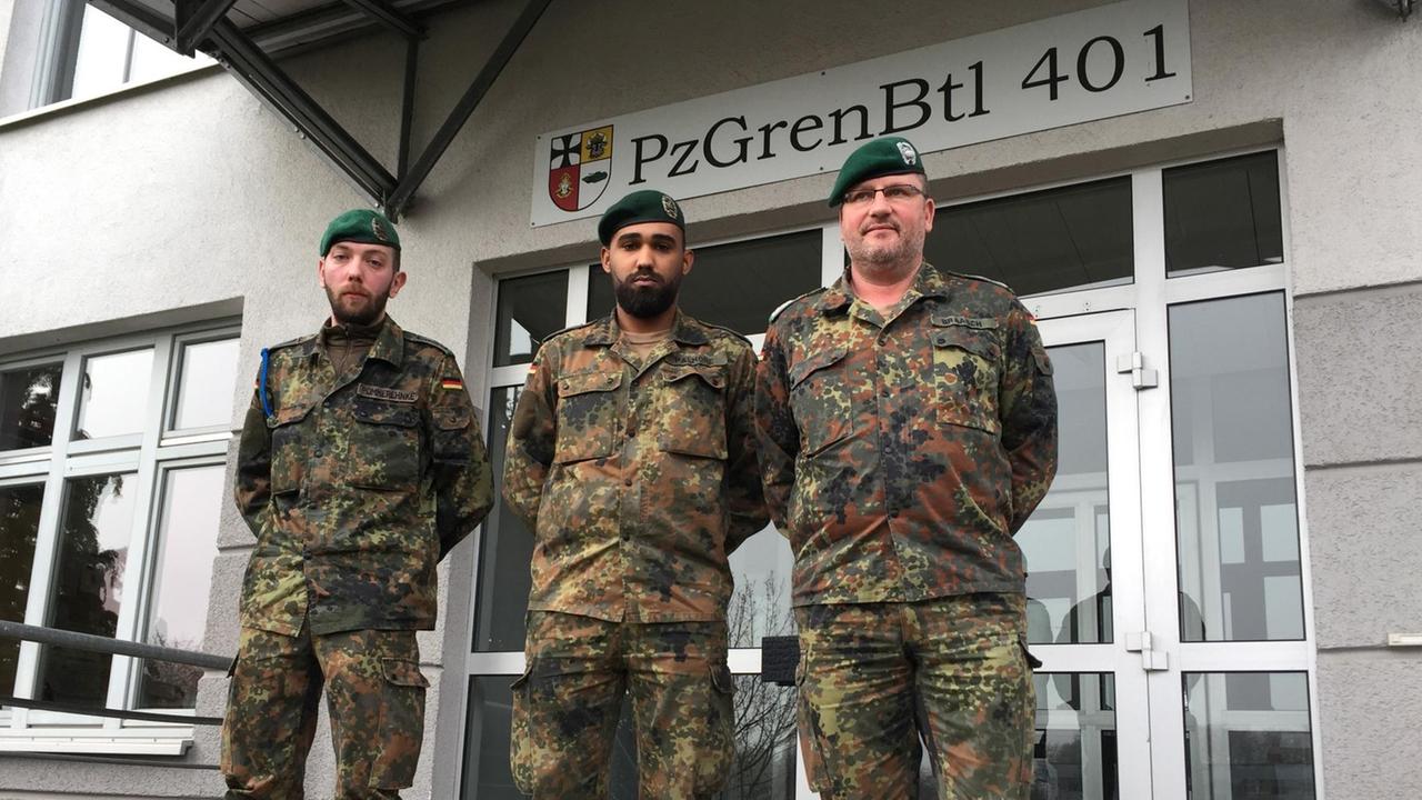 Hauptfeldwebel Jens Pommerehnke (l), Obergefreite Jerell Palmore und Stabsfeldwebel Stephan Braasch am Bundeswehrstandort in Hagenow. Drei Männer in Uniform stehen vor einem Gebäude.