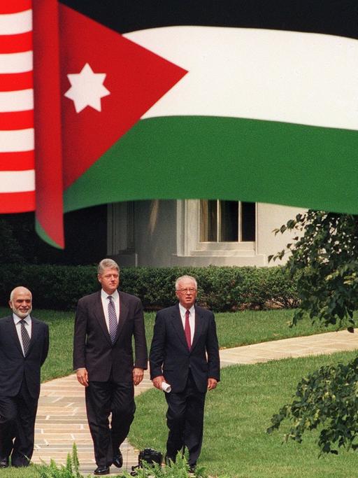 US-Präsident Bill Clinton (Mitte) mit Jordaniens König Hussein (links) und Israels Premier Yitzhak Rabin laufen einen Weg entlang, darüber hängen die Fahnen der drei Länder.