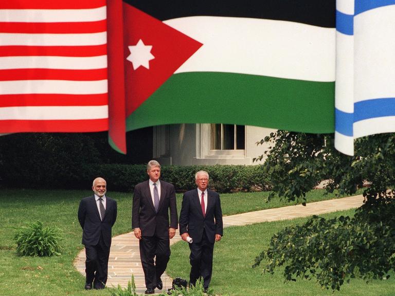 US-Präsident Bill Clinton (Mitte) mit Jordaniens König Hussein (links) und Israels Premier Yitzhak Rabin laufen einen Weg entlang, darüber hängen die Fahnen der drei Länder.