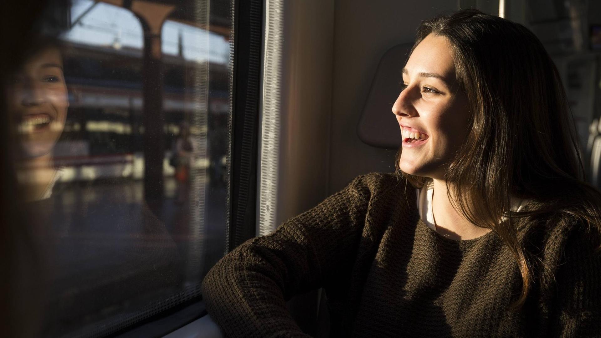 Eine fröhliche junge Frau sitzt in einem Zug und schaut aus dem Fenster auf den vorbeiziehenden Bahnhof. In der Scheibe spiegelt sich ihr lachendes Gesicht.