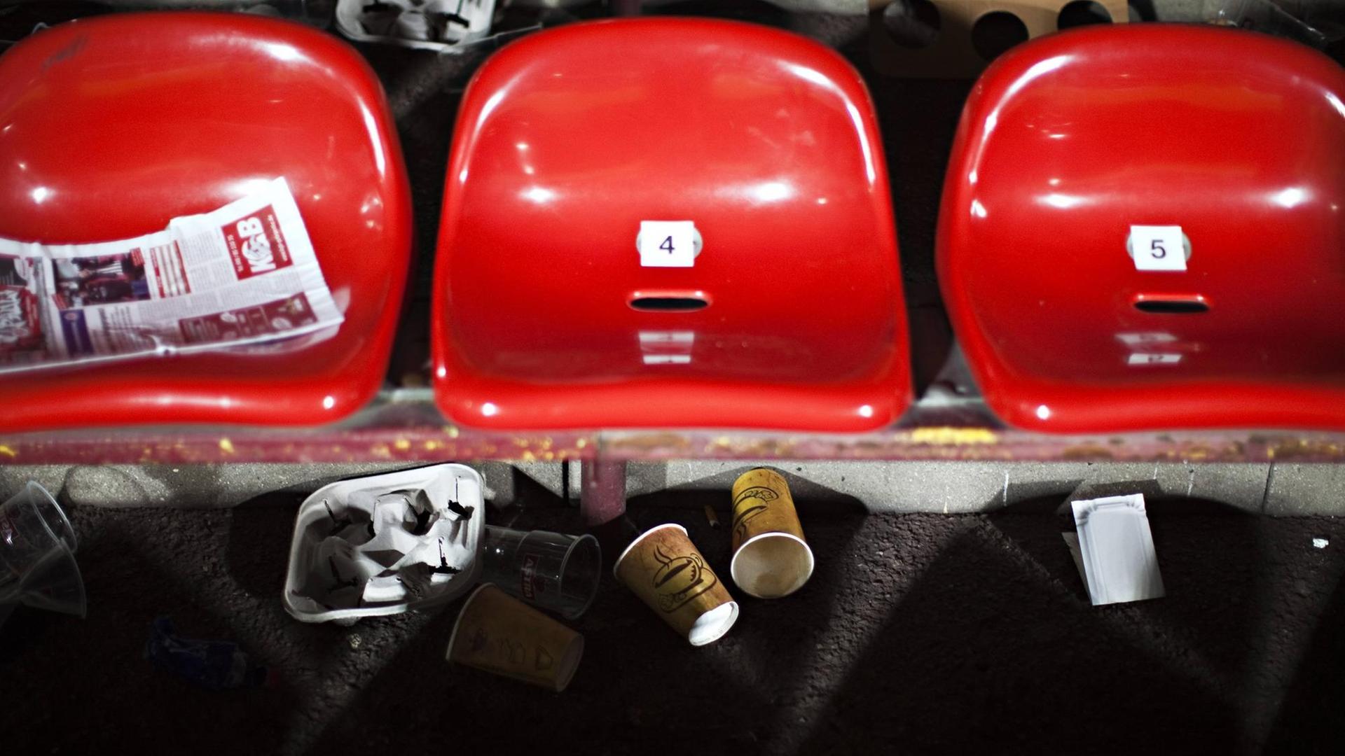Rote Sitzschalen einer Stadiontribüne mit liegengelassenen Papp- und Plastikbechern.