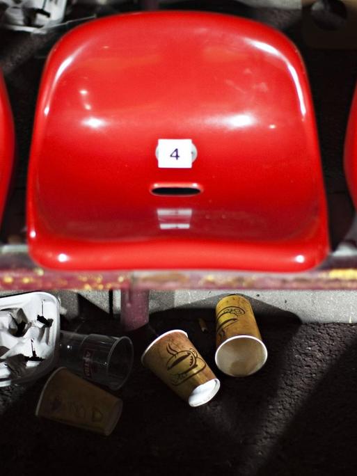 Rote Sitzschalen einer Stadiontribüne mit liegengelassenen Papp- und Plastikbechern.