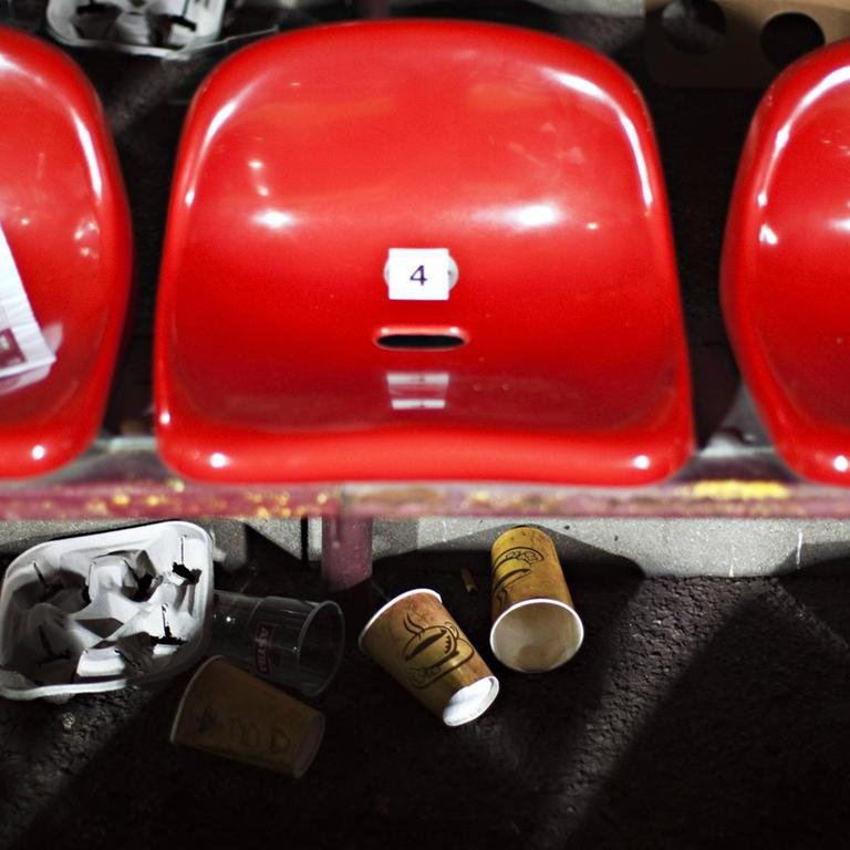 Rote Sitzschalen einer Stadiontribüne mit liegengelassenen Papp- und Plastikbechern.