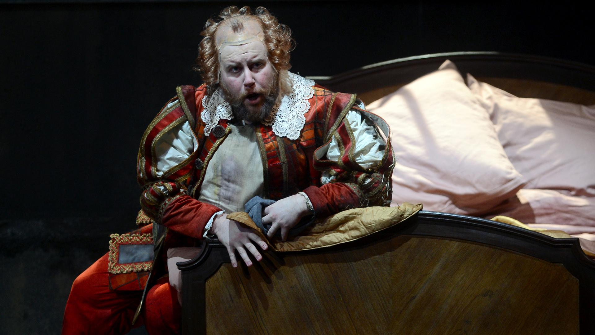 Der Sänger Noel Bouley sitzt kostümiert als Falstaff auf einem Bett.