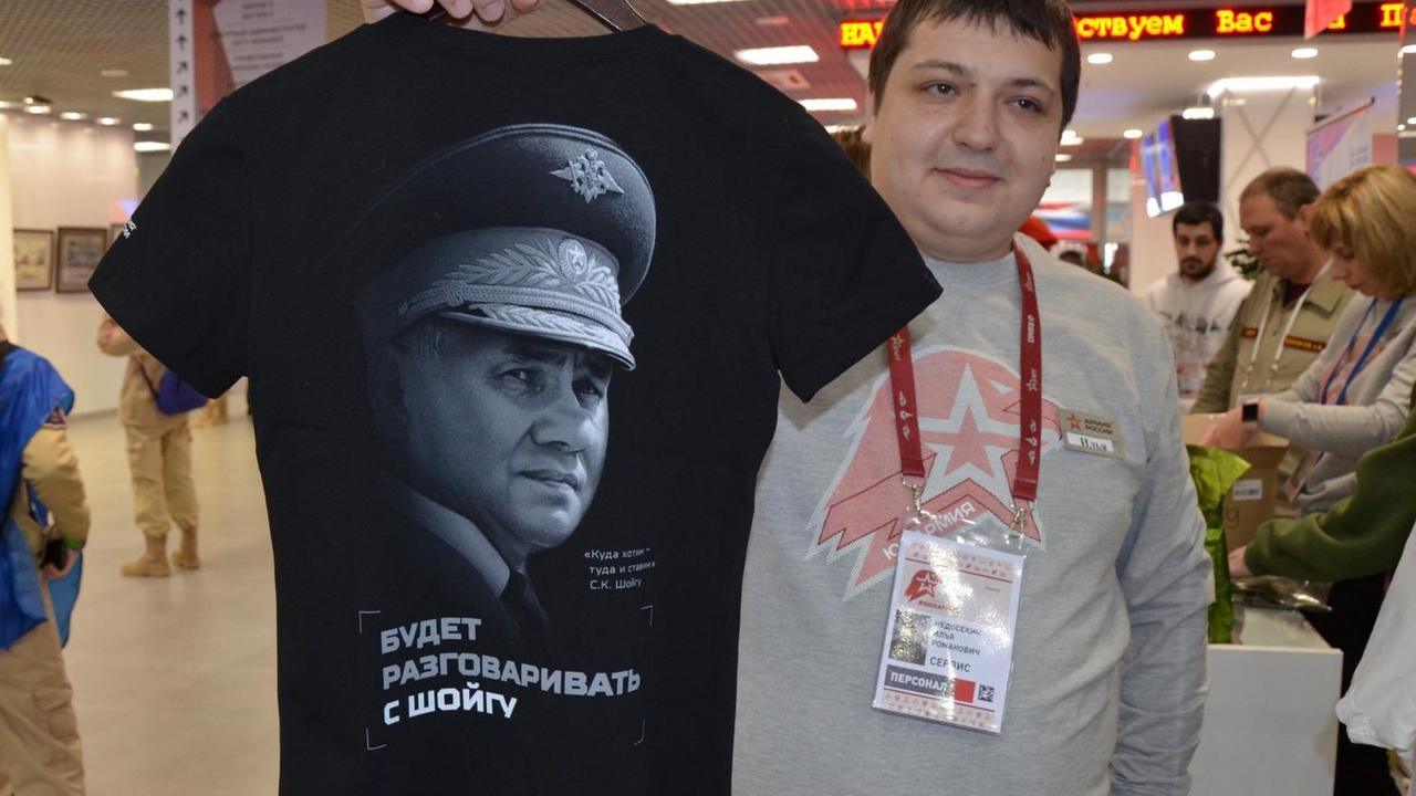 Ein Mann hält ein schwarzes T-Shirt in die Luft auf dem auf Russisch der Satz "... wird mit Schoigu sprechen" steht