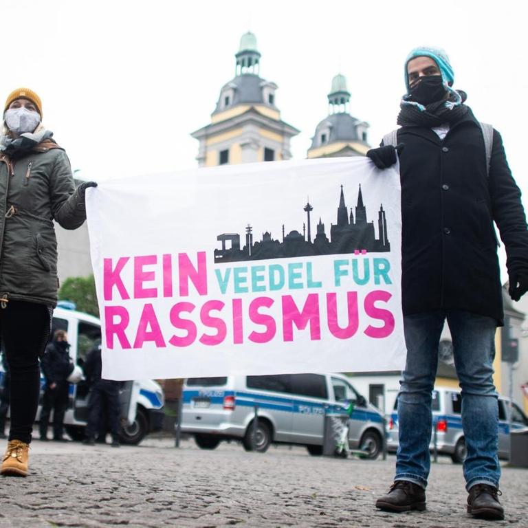 Zwei Demonstrierende mit Mund-Nasen-Schutz halten ein Plakat mit der Aufschrift „Kein Veedel für Rassismus“ hoch.