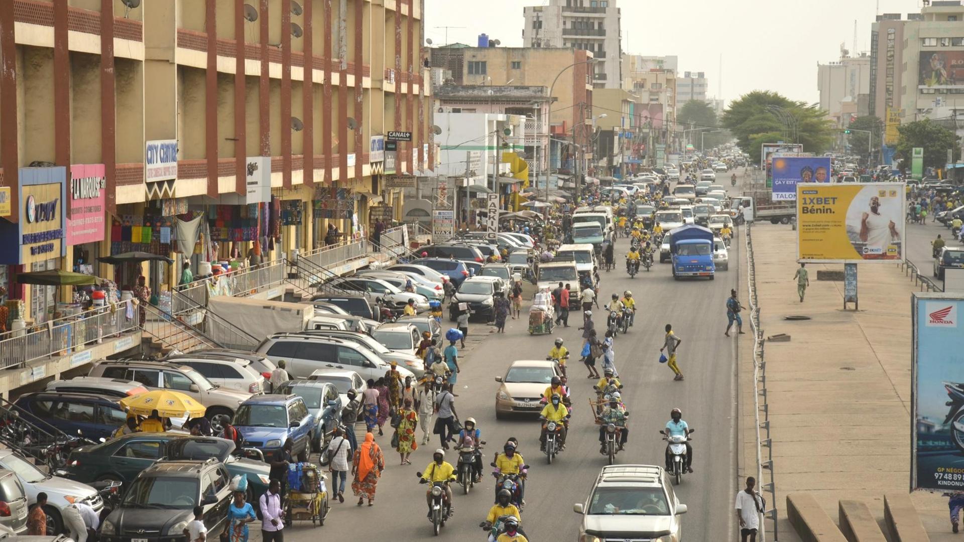Über eine große Straße in Cotonou rasen neben Menschen und Autos die typischen Zémidjans, Moped-Taxen, durch den dichten Verkehr.