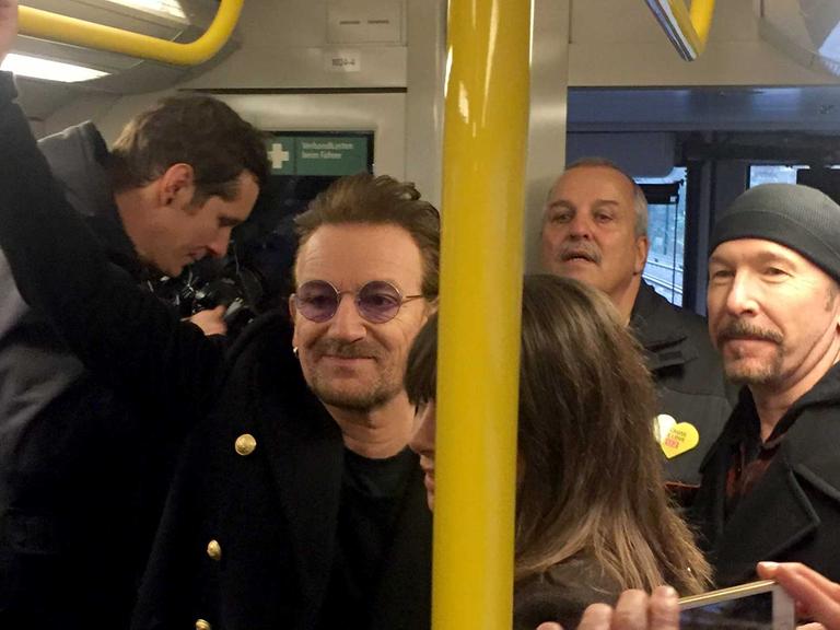Sänger Bono (l) und der Gitarrist David Howell Evans "The Edge" (r) von der Band U2 stehen am 06.12.2017 in der U-Bahn der Linie U2. Dicht gedrängt standen Fans und Medienvertreter in der U-Bahn, während die Musiker durch den Zug gingen, Hände schüttelten und Fragen beantworteten.