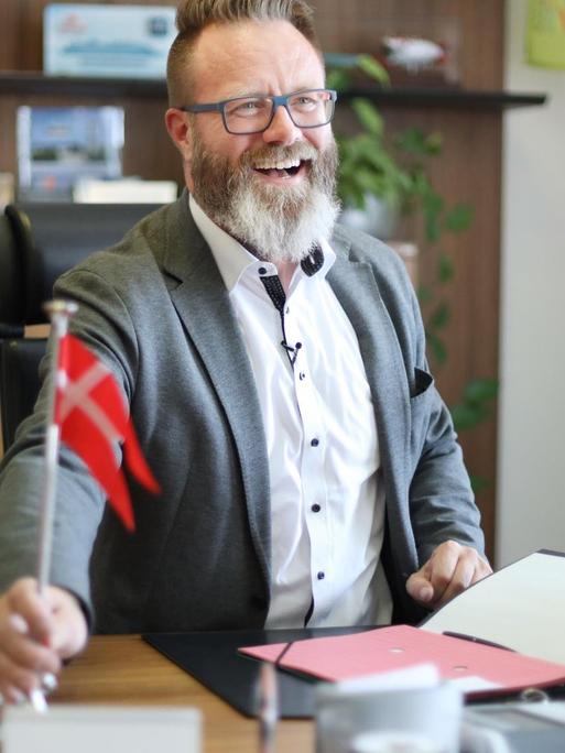 Rostocks neuer Oberbürgermeister Claus Ruhe Madsen sitzt am Schreibtisch seines Büro. Er lacht ind hält in seiner rechten Hand die dänische Flagge.